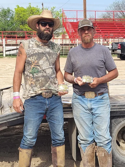 Two buckle winners, one in a straw cowboy hat, holding their prizes at the arena with red bleachers behind.