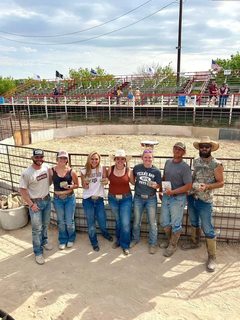 Seven buckle winners lined up in the arena ring, with flags flying over the red bleachers behind them.