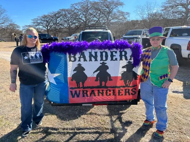 Two Bandera Wranglers members in parade costume standing beside the club's Texas-flag banner on a trailer.